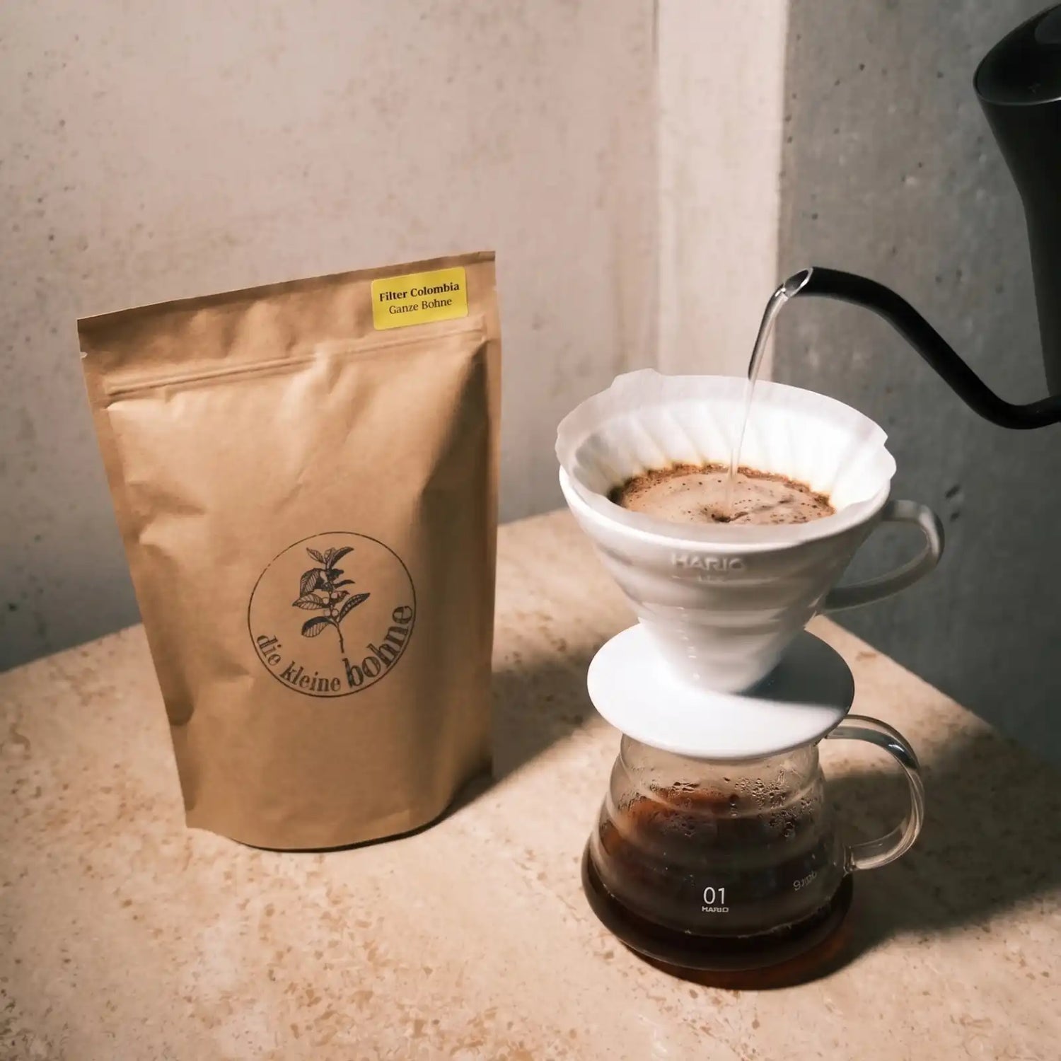 Bild einer Kaffeetüte Filter Colombia von diekleinebohne.de neben einem Hario V60 Handfilter, in den von einem Schwanenhalskessel heißes Wasser eingegossen wird.